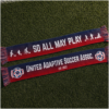 Two HD woven supporter scarves are laid almost flat on artificial soccer turf, displayed front and back. The scarf features a navy-to-red horizontal gradient with white tassels woven from navy, red, and white threads. One side reads "SO ALL MAY PLAY" in large white lettering, flanked by adaptive soccer icons representing blind soccer, intellectual disability soccer, frame soccer, amputee soccer, and power soccer. The reverse reads "UNITED ADAPTIVE SOCCER ASSOC." with "EST. 2023" beneath, and features the United Adaptive Soccer Association logo woven into both ends. The scarves are photographed in soft natural light, highlighting the woven texture and premium finish against the green turf background.