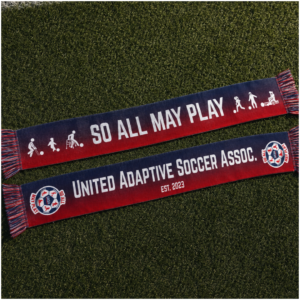 Two HD woven supporter scarves are laid almost flat on artificial soccer turf, displayed front and back. The scarf features a navy-to-red horizontal gradient with white tassels woven from navy, red, and white threads. One side reads "SO ALL MAY PLAY" in large white lettering, flanked by adaptive soccer icons representing blind soccer, intellectual disability soccer, frame soccer, amputee soccer, and power soccer. The reverse reads "UNITED ADAPTIVE SOCCER ASSOC." with "EST. 2023" beneath, and features the United Adaptive Soccer Association logo woven into both ends. The scarves are photographed in soft natural light, highlighting the woven texture and premium finish against the green turf background.