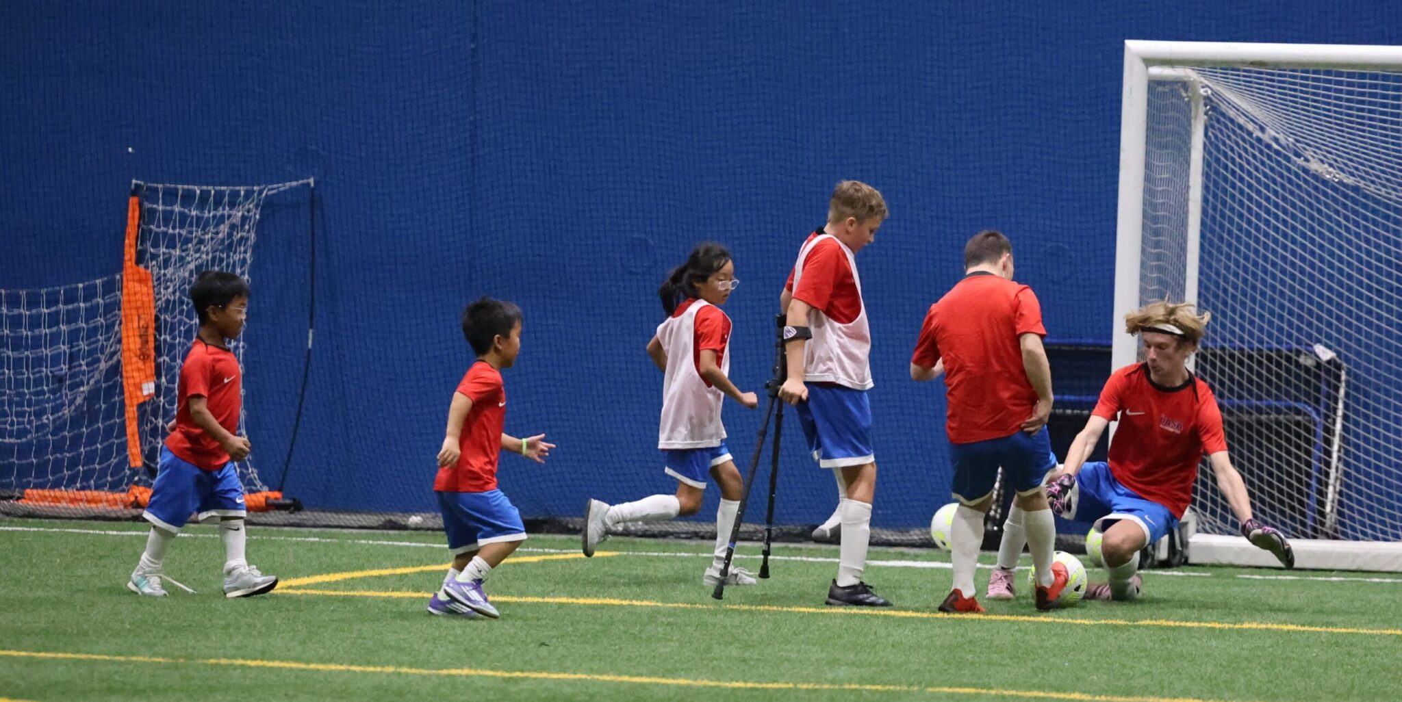 A group of young adpative soccer players of all disabilities take part in a scrimmage, as a goalkeeper makes a save and the other chase the rebound.