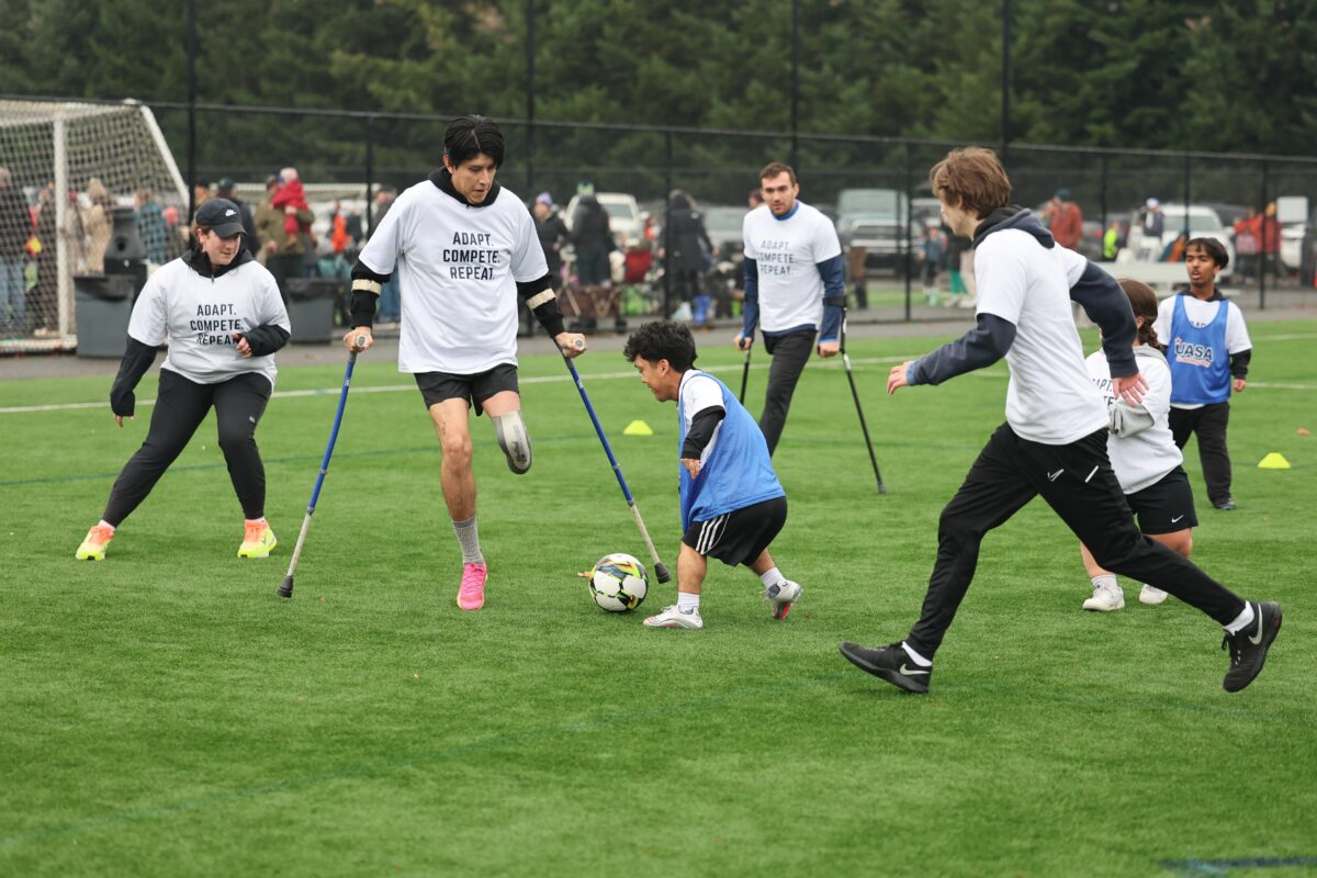 Action photo from an adaptive soccer clinic on an outdoor field. In the center, an amputee soccer player wearing a white "Adapt. Compete. Repeat." shirt, using forearm crutches and a lower-limb prosthesis, challenges a dwarf soccer player for the ball. Around them, several other athletes with a range of disabilities—including additional amputee players using crutches and players of short stature—take part in the small-sided game. Cones mark the playing area, while spectators, additional participants, and soccer goals are visible in the background, creating the atmosphere of a busy community soccer event.