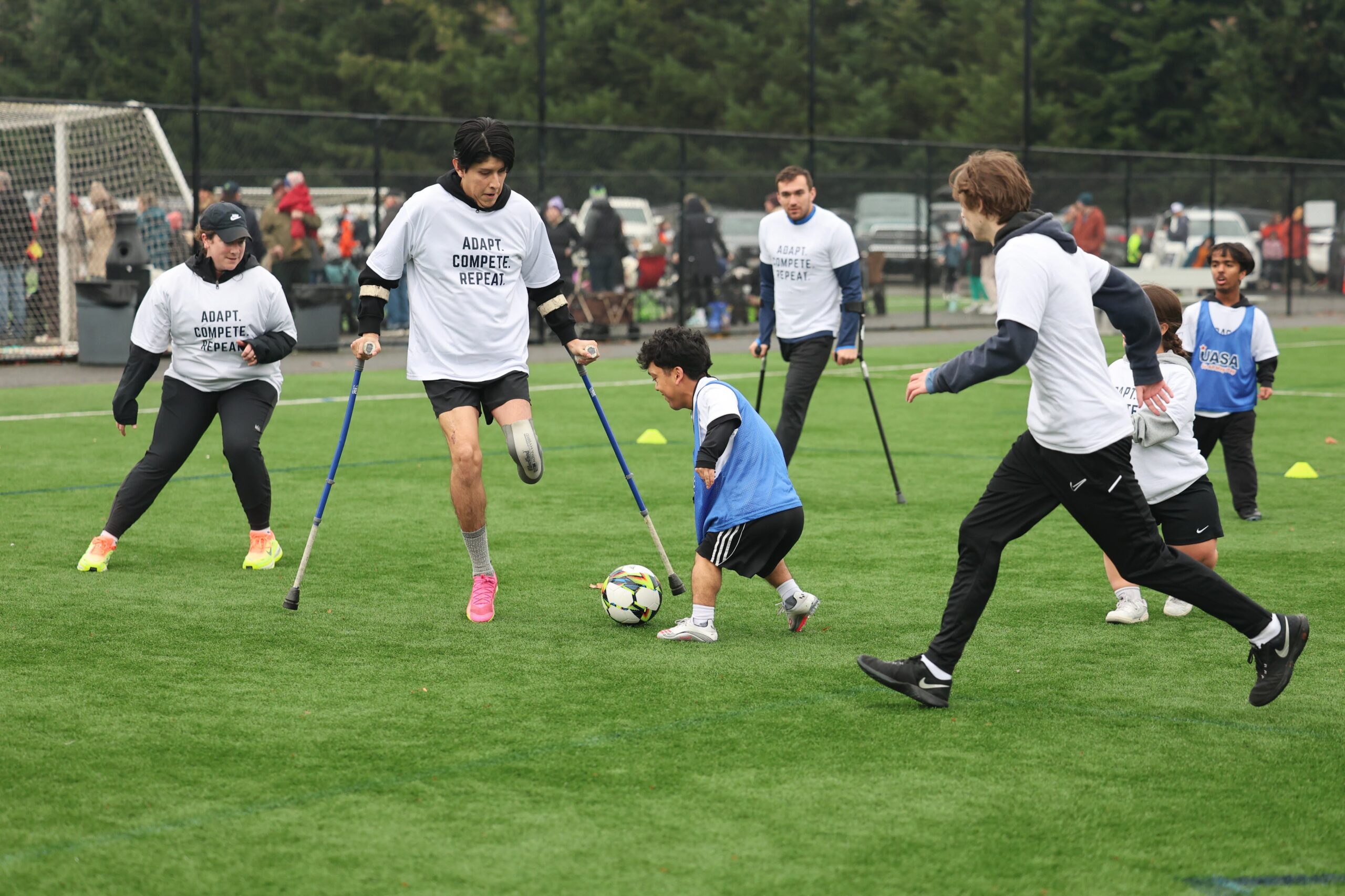 Action photo from an adaptive soccer clinic on an outdoor field. In the center, an amputee soccer player wearing a white "Adapt. Compete. Repeat." shirt, using forearm crutches and a lower-limb prosthesis, challenges a dwarf soccer player for the ball. Around them, several other athletes with a range of disabilities—including additional amputee players using crutches and players of short stature—take part in the small-sided game. Cones mark the playing area, while spectators, additional participants, and soccer goals are visible in the background, creating the atmosphere of a busy community soccer event.